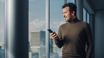 Thoughtful Arab Businessman Using Smartphone Standing in Office Looking out of the Window on Big...