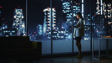 Fototapeta na wymiar Big City Modern Office at Night: Successful Young Businesswoman Standing and Using Laptop. Beautiful Female Digital Entrepreneur Thinking of Investment Strategy for e-Commerce Project.