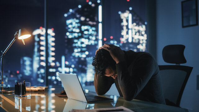 Portrait of Stressed Businessman Working on Laptop Computer in His Big City Office at Night. Overworked Tired Entrepreneur trying to Find Solution for Business Problems.