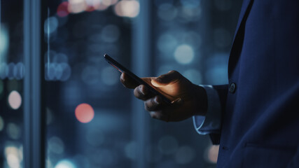 Night Office: Successful Black Businessman Wearing Suit Standing, Using Smartphone. CEO Browsing...