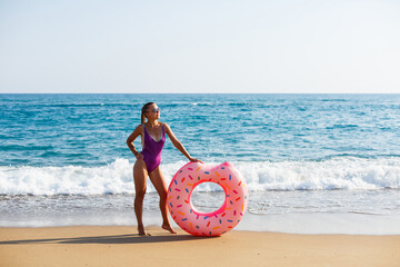 Naklejka na ściany i meble Attractive young woman in a bright swimsuit posing on the beach with an inflatable ring. Beautiful blond woman with long hair relaxing by the ocean. The concept of a sports model, swimwear