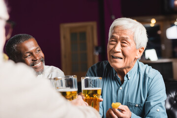 happy asian man holding beer and chips near blurred multiethnic friends in pub