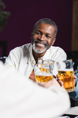 cheerful african american man clinking beer glasses with blurred senior friends in pub
