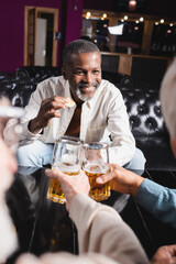 happy senior african american man holding chips while clinking glasses of beer with blurred friends