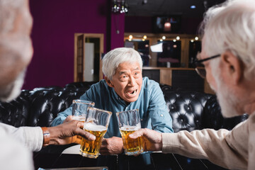 astonished asian man clinking glasses of beer with blurred interracial friends in pub