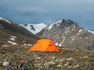 Orange tent on a sunny mountain slope. Camping on a rocky high-altitude plateau. Tent on the background of glacier and high snow-capped mountains. Solo trekking.