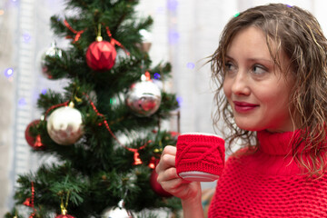 Young beautiful woman in a red sweater with a mug of cocoa and marshmallows at home by the new year tree on new year's eve. Selective focus. Portrait