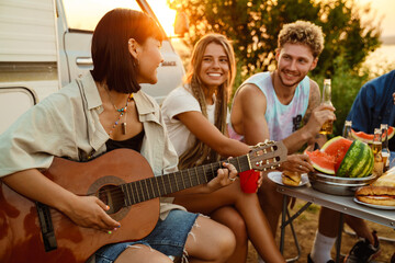 Multiracial friends smiling and playing guitar during picnic by trailer