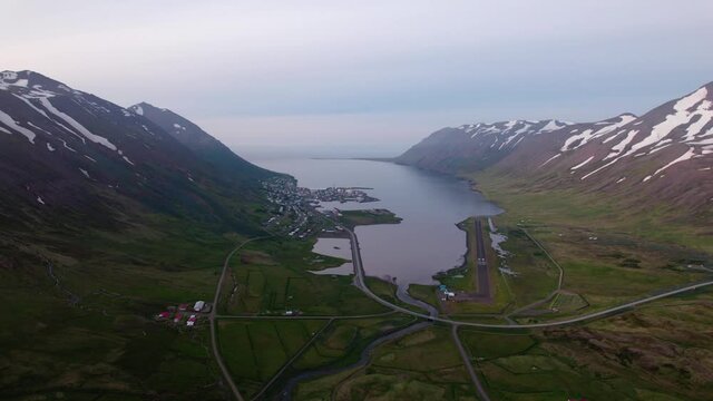 Aerial shot with view of coastal town in Iceland, surrounded by snowy mountains in a fjord.