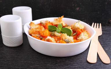 Pumpkin cut into pieces and baked with bread, dry herbs, olive oil and pumpkin seeds in a white plate on the black background.