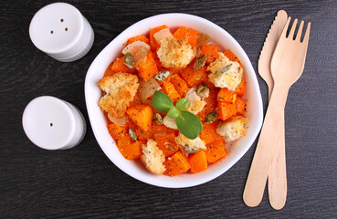 Pumpkin cut into pieces and baked with bread, dry herbs, olive oil and pumpkin seeds in a white plate, top view.