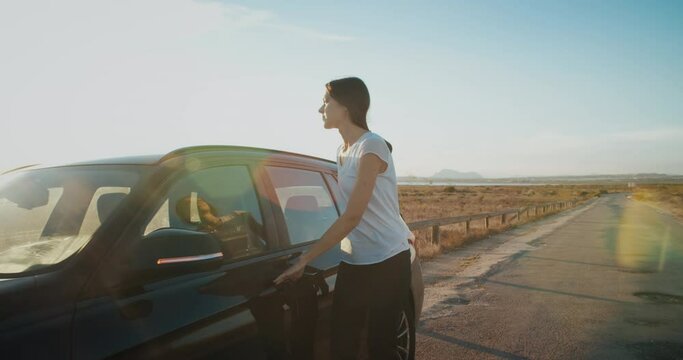 On a warm autumn evening in a clear sky on a country road, a girl closes the boot of a black car and walks into the sunlight to get behind the wheel of the car to continue her journey.