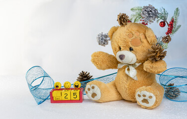 Christmas composition: a teddy bear juggles pine cones, a festive blue lace ribbon with sequins, a voluminous toy calendar with funny faces on the background of a New Year's wreath. Selective focus