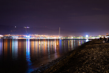 Night city with lights and mountains by the sea and red sky, pebble shore.