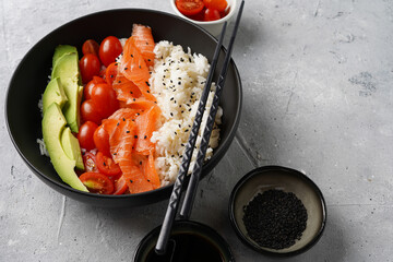 Poke bowl - black bowl with white rice, smoked salmon, cherry tomatoes and avocado, sesame seeds, soy sauce and chopsticks on a grey concrete surface