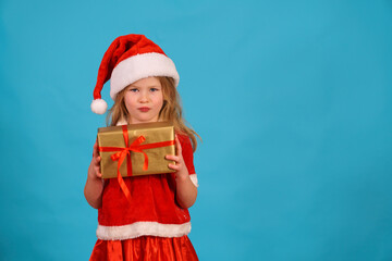 Funny Christmas child. girl in santa claus cap is holding gift and grimacing. Play with facial expressions. Sly look of satisfied girl with gift. Mischievous and naughty with gift on blue background.