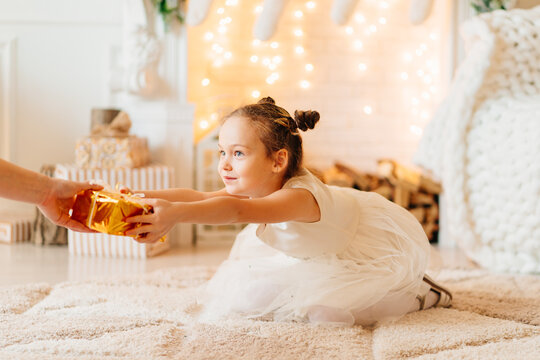 A Little Girl In A White Dress Sits On The Floor On A Rug Receives A Gift In Gold Wrapping Paper. Tradition To Give Gifts To Children For The New Year And Birthday. Santa Claus. Children's Toy Store.