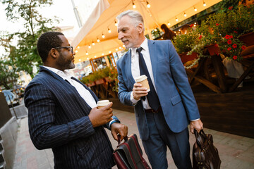 Multiracial two men wearing suits talking while drinking coffee