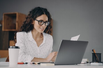 Happy young european woman wearing glasses in white shirt working on modern laptop from home