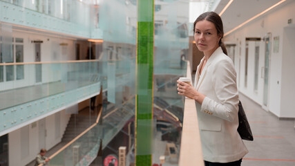 Fototapeta premium young business woman in a white jacket is standing in a public space with a takeaway coffee in her hand. Confident brunette freelancer, portrait on the background of a business center.