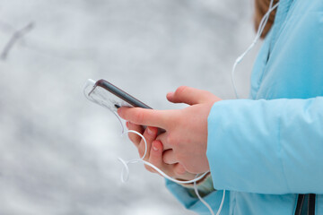 Close up shot of frozen frostbitten hands of a young pretty girl who walks in a winter snow park watching videos online in a smartphone or listening to music with headphones