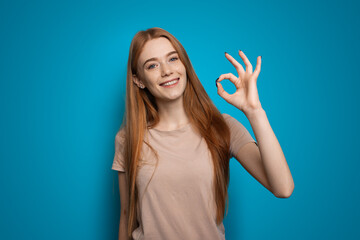 Fototapeta premium Portrait of smiling redhead woman looking at camera, showing okay sign standing over blue background. No problem gesture.
