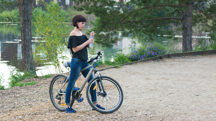 Fototapeta premium A brunette woman on a bicycle stopped by river to drink water from bottle.