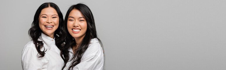 pleased asian mother and young daughter in white shirts hugging isolated on grey, banner