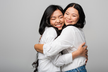pleased asian mother and daughter in white shirts hugging isolated on grey