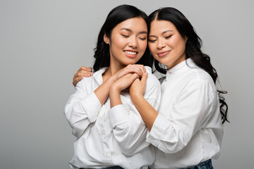 cheerful asian mother and young adult daughter hugging each other and holding hands isolated on grey