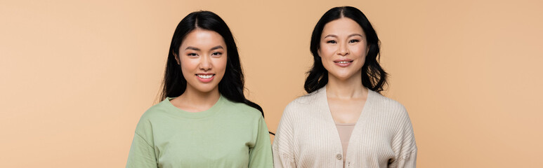 cheerful asian mother and adult daughter looking at camera isolated on beige, banner