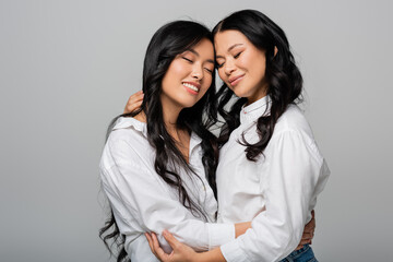 joyful asian mother and young daughter in white shirts embracing isolated on grey