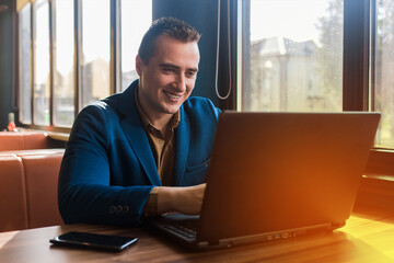 A business smiling man happy businessman a stylish portrait of Caucasian appearance in a jacket works in a laptop or computer, sitting at a table by the window in a cafe