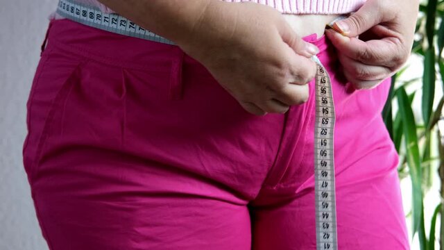 fat woman in pink sweater measures waist circumference with centimeter tape, belly stands, concept time to lose weight, wrong lifestyle, consequences of inactivity, unhealthy diet