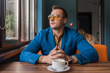 A brooding businessman a stylish portrait of Caucasian appearance in sunglasses, sits idly by at a table on a coffee break in a cafe and looks out the window