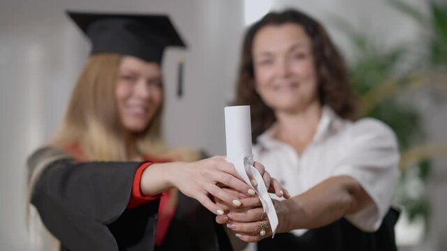 Close-up rolled graduation diploma with ribbon in hands of happy Caucasian mother and daughter. Proud mature and young women holding certificate standing at home indoors