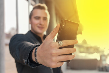 A young satisfied guy holds a smartphone or mobile phone in his hand, takes a selfie or talks on video on the outdoor street, close-up