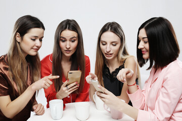 Four beautiful girlfriends in pajamas are sitting at the table, drinking tea or coffee and chatting in a social network.