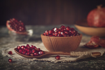 Pomegranate seeds lying in a wooden spoon, and in a wooden plate on a linen napkin. In the background are pomegranates with a slice of pomegranate.