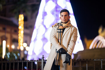 Young man adjusting his gloves at night with scarf in winter. Christmas tree in the background.