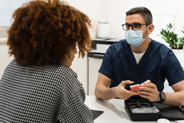Male dentist with dentures explaining to female patient in medical clinic during pandemic
