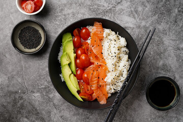 Poke bowl - black bowl with white rice, smoked salmon, cherry tomatoes and avocado, sesame seeds, soy sauce and chopsticks on a grey concrete surface, top view