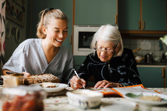 Senior woman solving crossword puzzle in book sitting by smiling nurse in kitchen at home