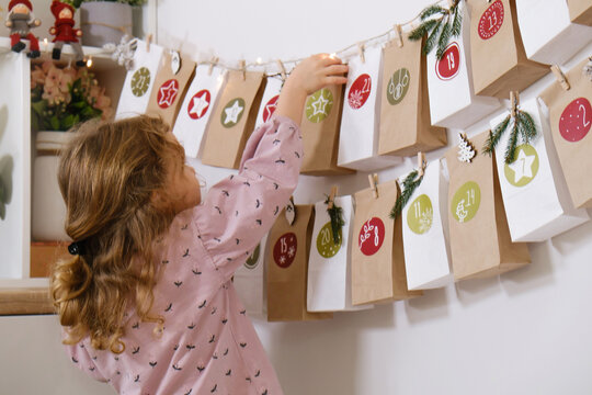 Toddler child gets new advent calendar task, waiting for Christmas. Festive mood for cute curly girl. Kid takes advent bag with dates from the wall at home.