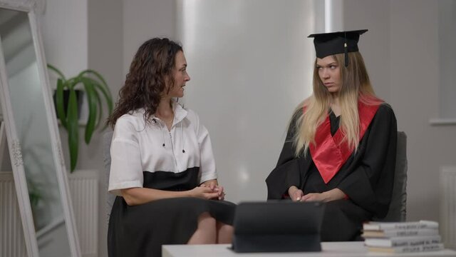 Serious Caucasian mature mother talking with young graduate daughter sitting indoors at home. Beautiful high school grad student with strict parent on graduation day. Family and lifestyle