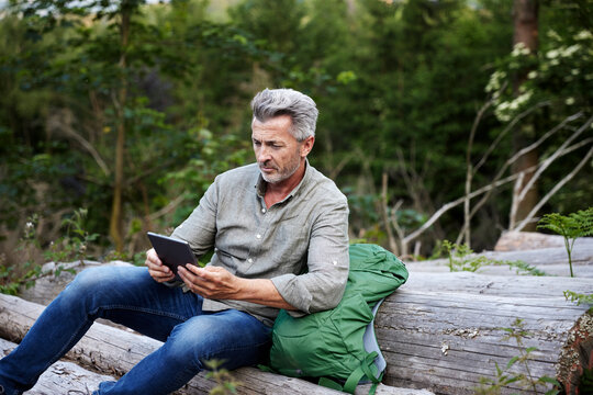 Male Hiker Using Digital Tablet While Sitting On Tree Trunk