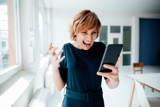 Excited businesswoman using mobile phone while standing in office - Powered by Adobe