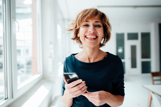 Cheerful businesswoman holding mobile phone while standing in office