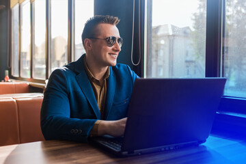 A business man happy smiling businessman a stylish portrait of Caucasian appearance in a jacket and shirt, sunglasses works in a laptop or computer, sitting at a table by the window in a cafe