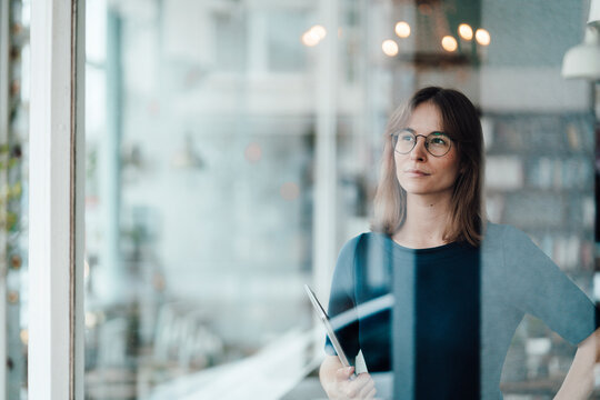 Young female professional wearing eyeglasses standing with laptop seen through glass window